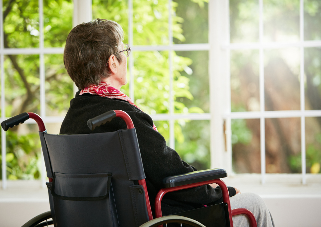 A senior woman sitting in a wheelchair looks through a window into a green, sunlit garden, frustrated that she cannot reach it.